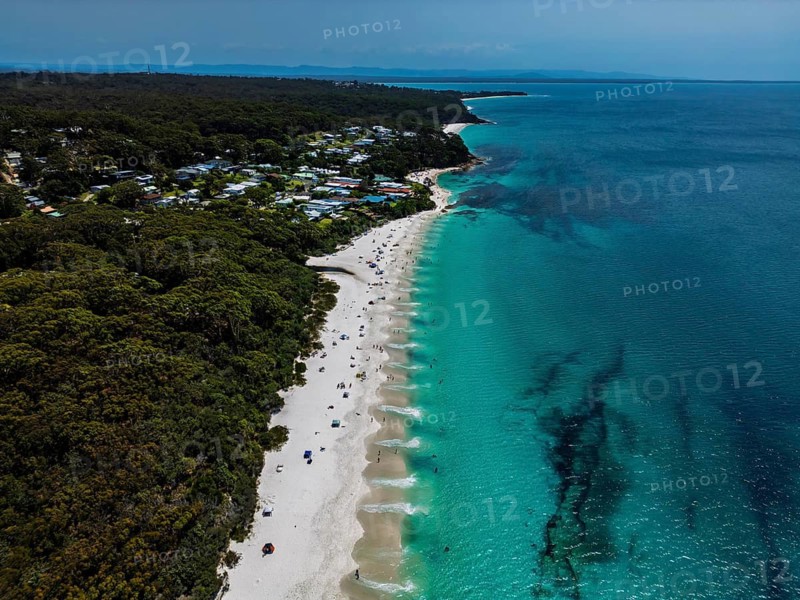 Aerial view of a peaceful beach with turquoise waters and lush forest surrounding a small coastal town in Jervis bay, Australia. The sandy shoreline is lively with people enjoying the sun
