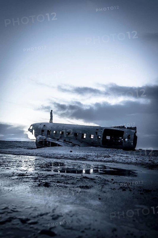 A man stands on a beach next to a large, old airplane that has crashed into the water