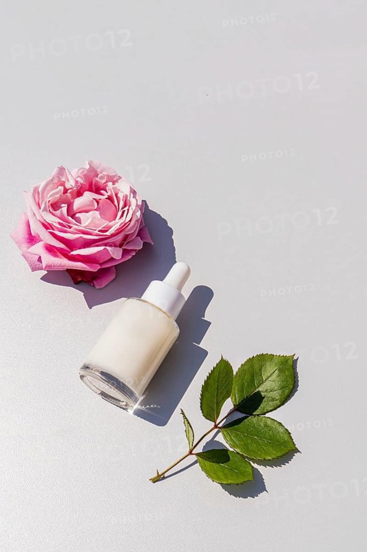 Top view of a cosmetic serum bottle with a white dropper, placed beside a pink rose and green leaves on a smooth, light-colored surface, creating a minimalist aesthetic