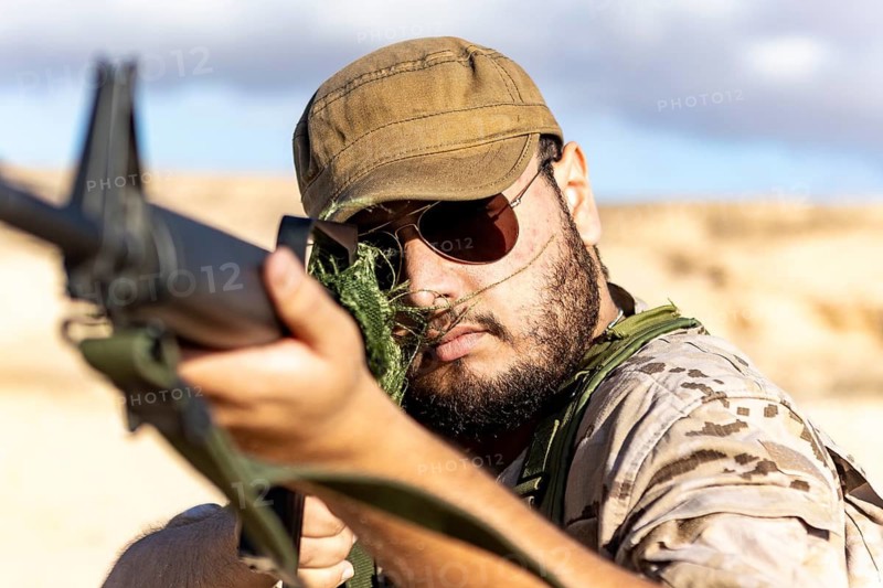 A soldier in camouflage uniform aiming a rifle in a desert landscape. The figure wears sunglasses and a cap, with a focused expression, conveying determination and focus