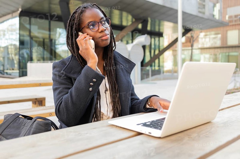 An African American businesswoman with braids is engaged in work using her laptop while taking a call on her mobile phone, seated outdoors