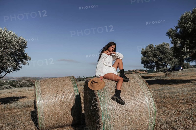 A young woman in a white dress sits atop hay bales in the Italian countryside, surrounded by olive trees under a clear blue sky, capturing the essence of serene rural life