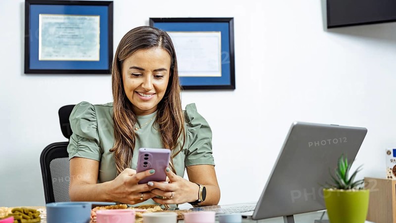 A nutritionist sits at her desk, smiling while using a smartphone, with a laptop open. Certificates on the wall indicate her professional qualifications