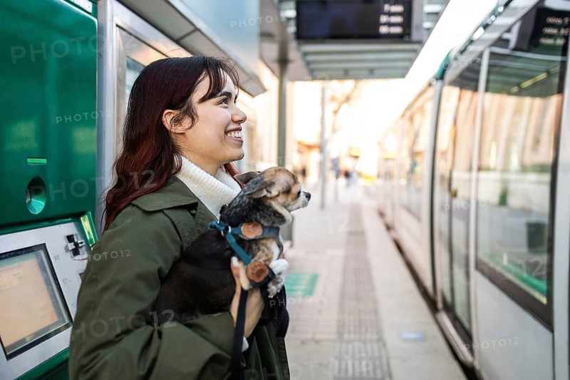 Smiling woman holding a small chihuahua at a tram station, excited to board a tram. The scene captures warmth, companionship, travel enthusiasm, and the joy of shared adventures