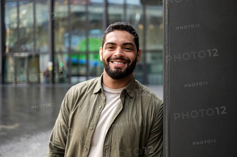 A confident man smiles warmly, wearing casual attire, while leaning against a wall in front of a modern building. His relaxed demeanor adds a welcoming touch to the urban setting