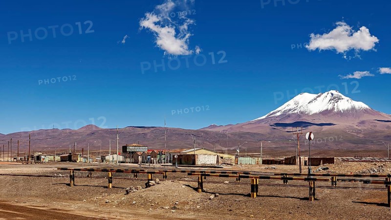 Scattered buildings under a snow-covered volcano in a dry landscape with clear skies, the town of Ollagüe in the Chilean Atacama Desert with the Ollagüe volcano of the same name in the background
