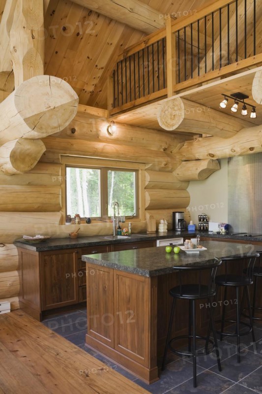 Island with dark grey granite countertop and black stained wooden bar stools in kitchen with ceramic tile floor inside handcrafted Eastern white pine Scandinavian log cabin home, Quebec, Canada