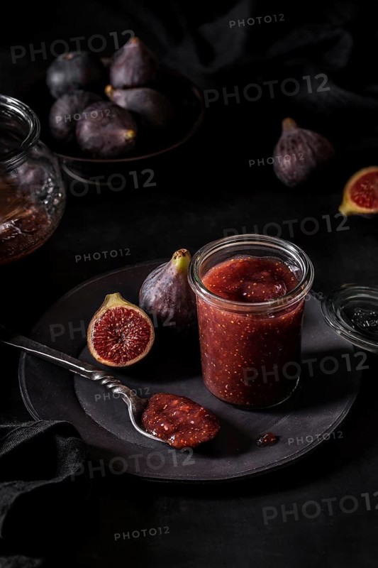 From above fresh fruit jam in small jar and on spoon with full and cut figs on plate at black kitchen table
