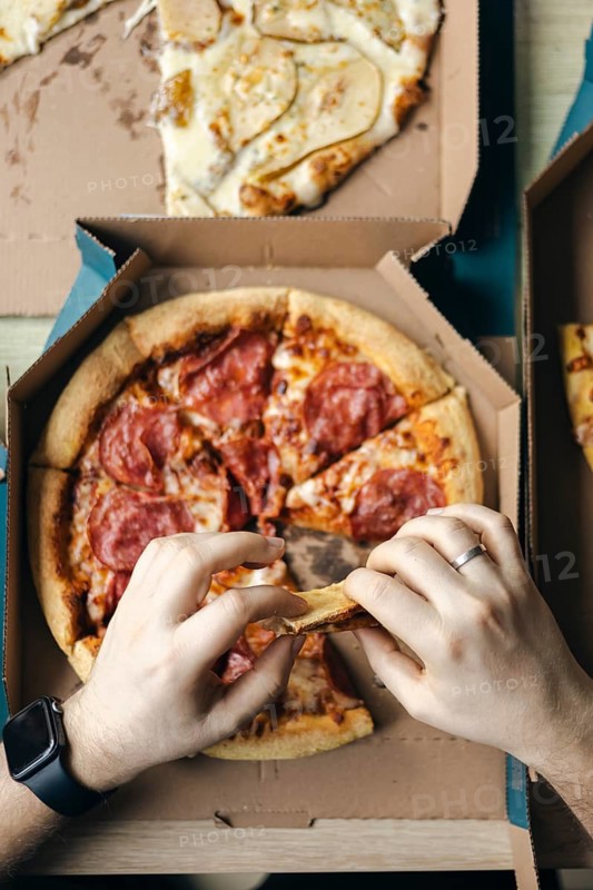 From above cropped unrecognizable hand taking slices of assorted pizzas from cardboard boxes, highlighting a casual social eating scenario indoors with a visible smartwatch worn by one person