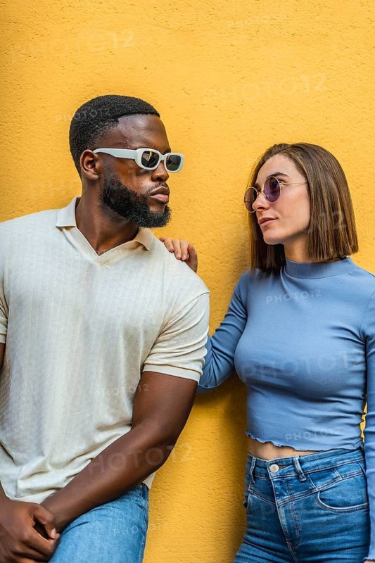 Vertical photo of a cool modern stylish multi-ethnic couple with denim clothes and futuristic sunglasses against yellow background int he city