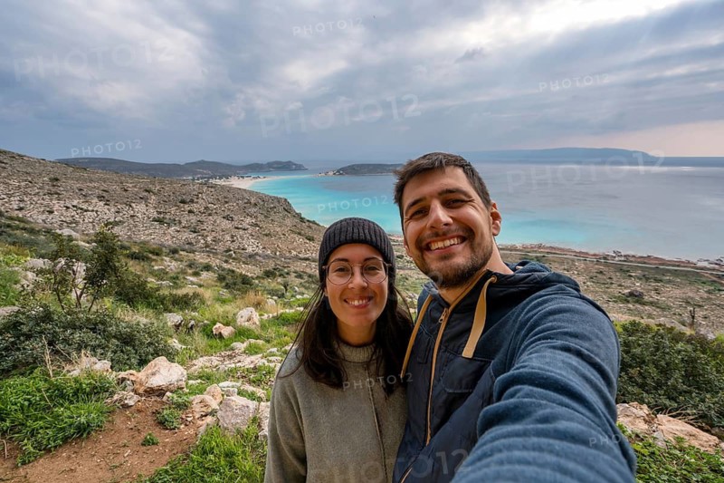 Happy couple taking a selfie with a stunning coastal backdrop in Greece. The vibrant turquoise sea, lush greenery, and cloudy sky create a picturesque setting