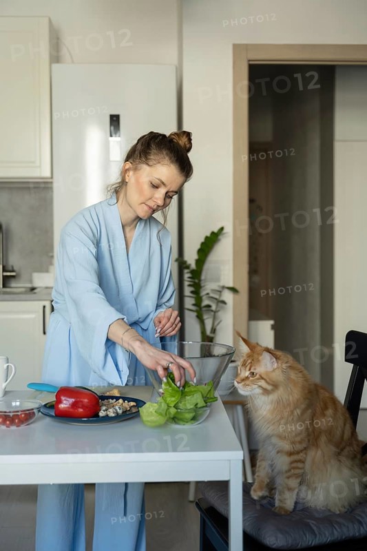 A woman in a blue robe prepares vegetables at a kitchen table. A curious cat sits nearby, observing her actions. The scene captures a cozy, domestic moment