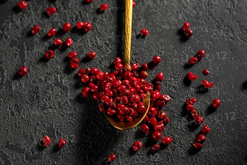 Top view of bright red peppercorns scattered from a golden spoon against a textured dark background, showcasing vivid color and texture contrasts in a gourmet setting