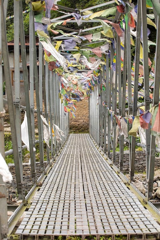 A suspension bridge on the Everest Trek is beautifully adorned with colorful prayer flags. These flags add a vibrant touch against the backdrop of lush greenery and rustic buildings