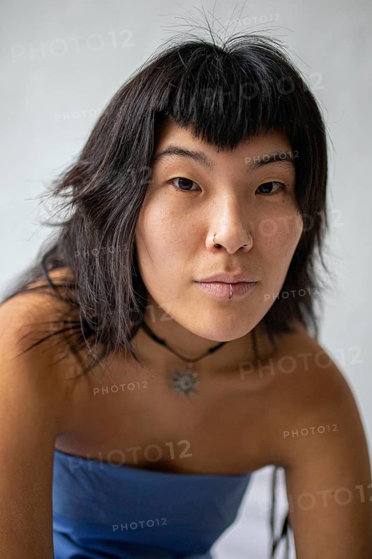A close-up portrait of a young Asian woman with dark hair, bangs, and facial piercings. She wears a choker necklace and a strapless blue top, gazing directly into the camera with confidence