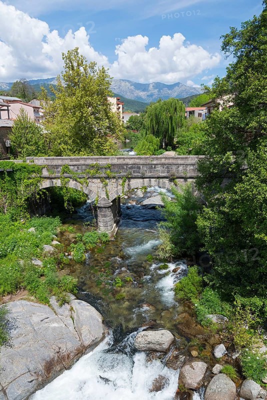 Eine Steinbrücke spannt sich über einen Fluss in einer malerischen grünen Landschaft unter blauem Himmel mit Bergen im Hintergrund, Arenas de San Pedro, Fluss Arenal, Ávila, Avila, Kastilien-León, Kastilien-Leon, Spanien