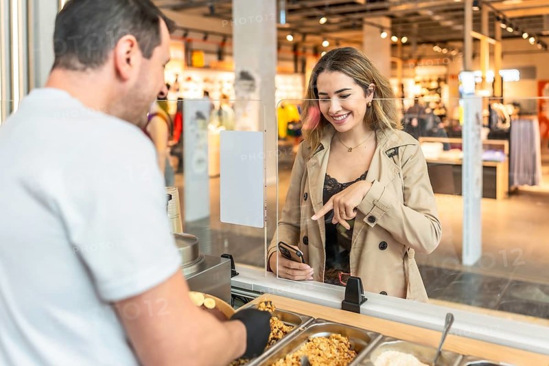 Young woman smiling and pointing at her smartphone screen while ordering a healthy meal from a male employee at a food counter in a bustling shopping center