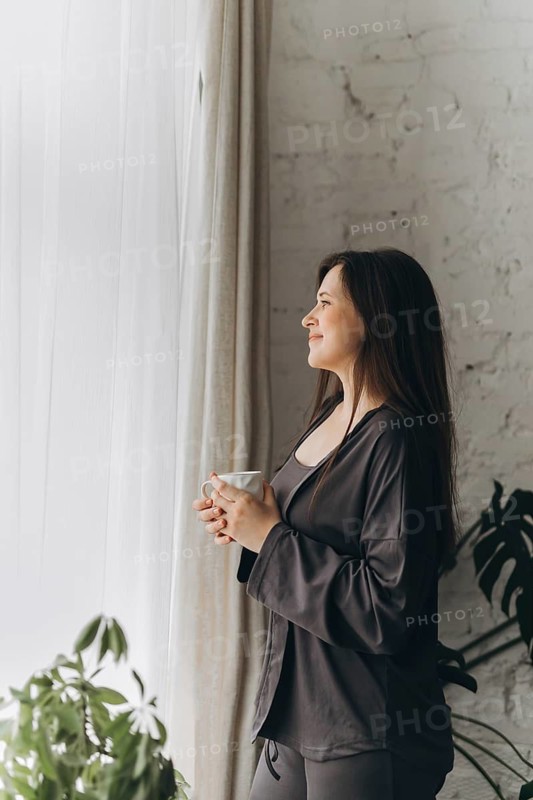 A woman stands by a window with a cup of coffee, enjoying the morning sunlight. Her serene expression suggests a moment of mindfulness in a cozy home setting