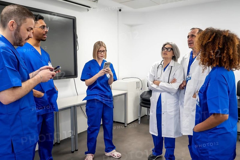 Healthcare professionals and medical students are engaging in a training session or meeting inside a modern hospital office, discussing information and using mobile devices to look up details