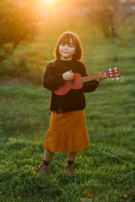 A young girl stands in a sunlit field, playing a ukulele. She wears a cozy sweater and mustard skirt, illuminated by the warm glow of the setting sun, creating a serene ambiance