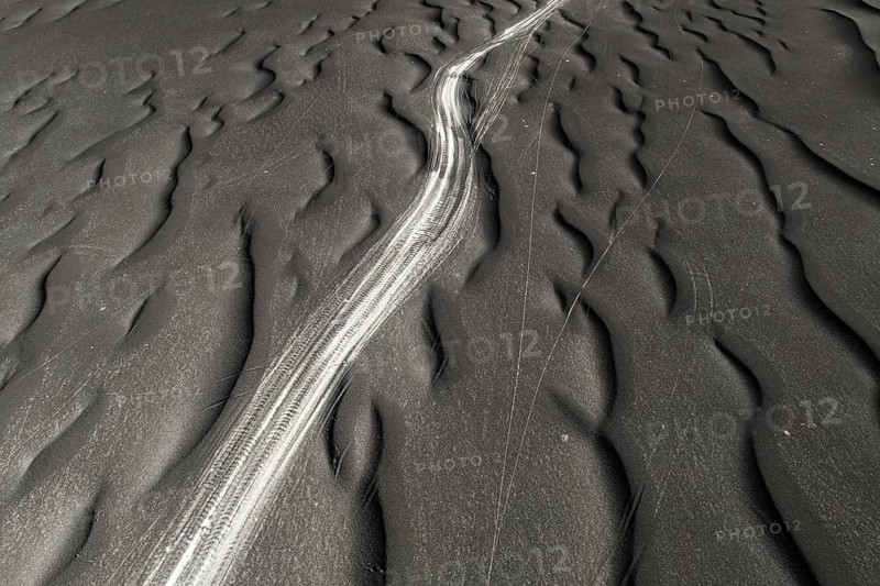 A striking image capturing the unique textures of black sand, highlighted by natural ripple patterns and a contrasting shiny trail possibly from a vehicle
