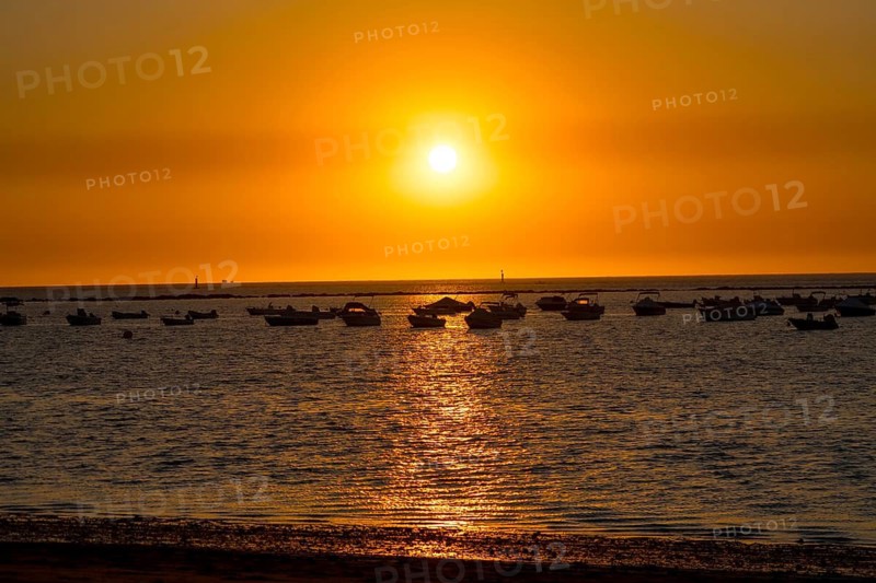 Several boats are silhouetted against a vibrant orange sunset over the ocean in sanlucar de barrameda, spain, creating a serene and picturesque scene