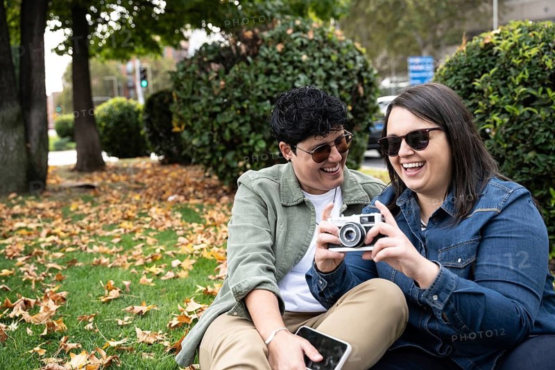 A cheerful curvy couple enjoys a day in the park, sharing moments and taking photos with a vintage camera. Surrounded by greenery and autumn leaves, they smile warmly