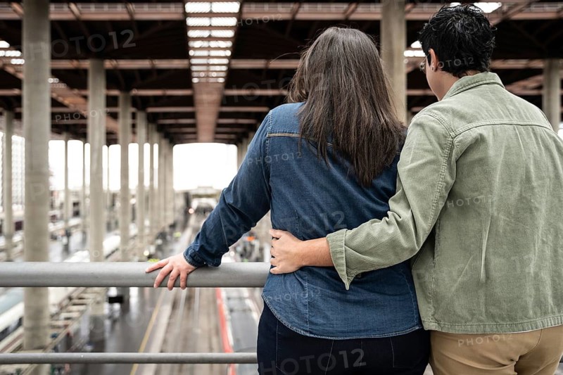 A loving lesbian couple embraces while standing on a balcony, overlooking a train station. Their relaxed posture and casual attire reflect a serene moment together