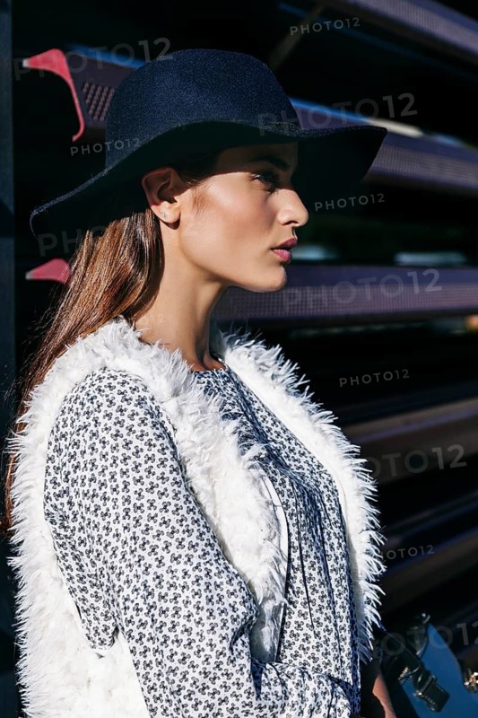 Woman in a black hat and white fur vest poses stylishly. Her patterned dress adds elegance, while the sun casts a soft glow on her confident expression