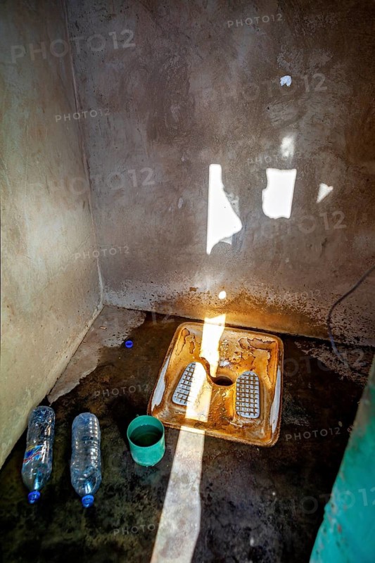 A shadowy bathroom with a simple toilet, two water bottles and a cup. Light falls through a window, view into a public toilet in the Sahara in Algeria