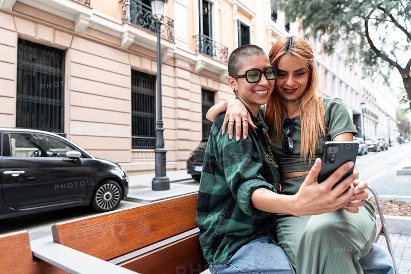 A happy Colombian lesbian couple takes a selfie while sitting on a bench in an urban setting, celebrating love, diversity, and LGBTQIA+ pride with joy and authenticity