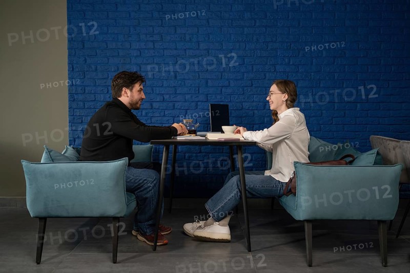 Two people are seated at a table in a cozy cafe, engaged in conversation over tea. They appear relaxed, with notebooks and a laptop on the table. The setting is casual yet professional