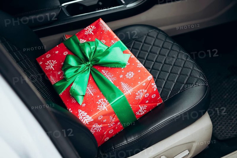A vibrant red gift box wrapped with festive patterns and a green ribbon sits on a car seat, capturing a moment of holiday excitement and surprise during a journey