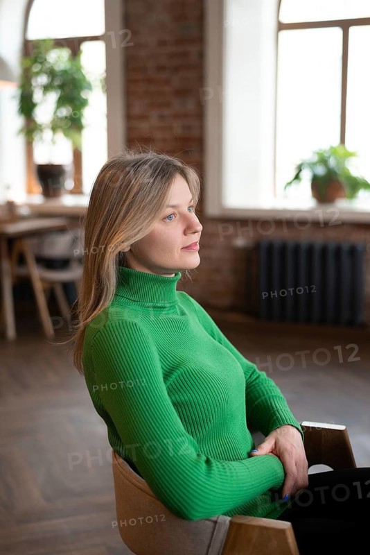 A young woman in a green sweater sits alone in a cozy cafe by the window, gazing thoughtfully outside. The warm, natural light and relaxed ambiance create a peaceful atmosphere