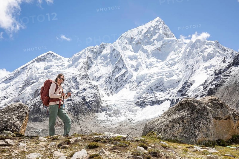 A woman treks in the Himalayas, with snow-capped Everest in the background. She is equipped with a backpack and trekking poles, enjoying the breathtaking mountain scenery