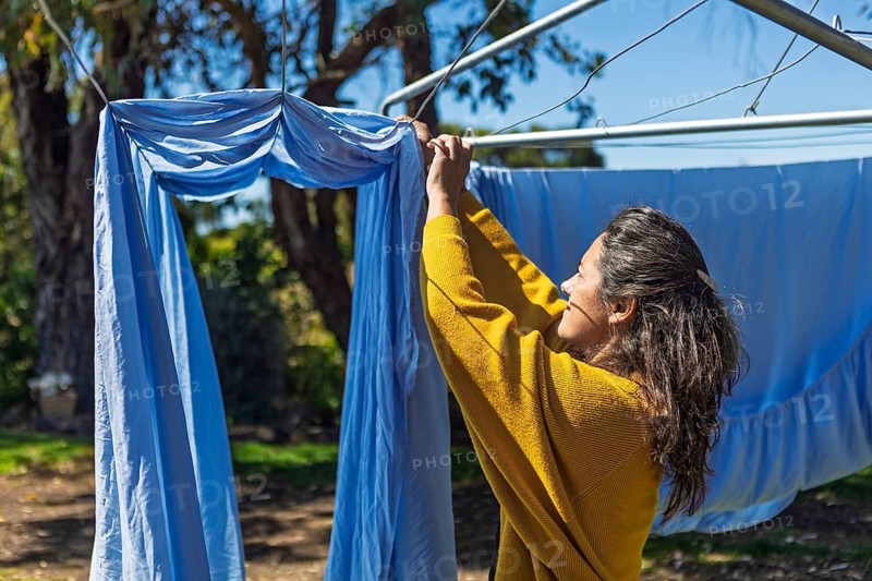 A woman hangs fresh blue sheets on a clothesline in the backyard, capturing daily routines in an Australian home setting under clear skies