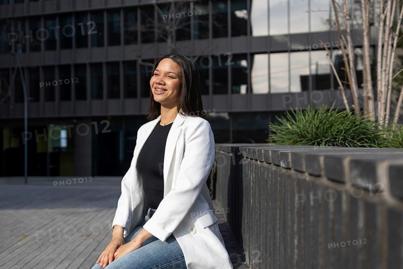 A confident woman wearing a white blazer sits outdoors, smiling in an urban setting. The modern building background reflects a professional business lifestyle theme