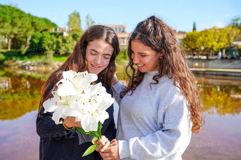 A young romantic LGTBIQ+ couple enjoys a sunny day outdoors while holding and admiring a large bouquet of white lilies. Both are looking at the flowers, creating a candid moment of connection and affection