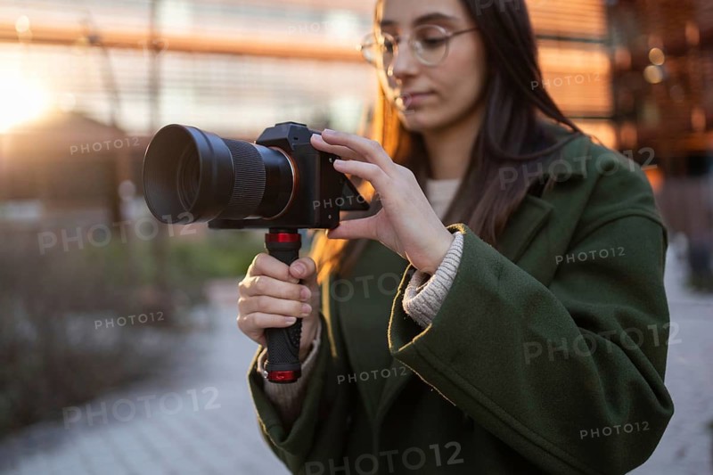 A woman in a green coat holds a camera while outdoors in a sunset setting The light softly illuminates her focused expression, capturing a moment of concentration