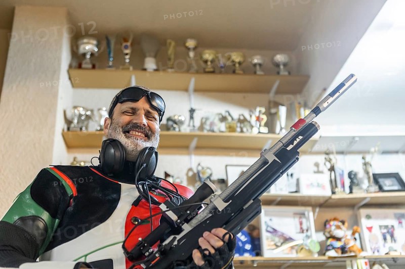 Blind athlete proudly holds a rifle, surrounded by numerous trophies and awards on shelves, symbolizing achievement and excellence in blind Olympic shooting sports