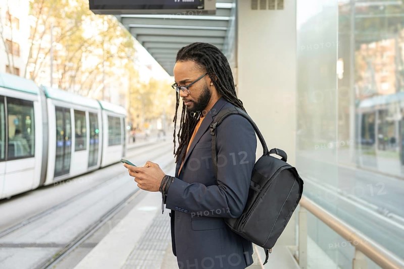 A cuban businessman in a suit, carrying a backpack, uses his phone while waiting at a tram stop. Captures the essence of modern urban life and technology in daily commutes