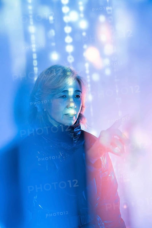 An elderly Asian woman interacts with a digital interface within the metaverse. She is surrounded by vibrant, glowing lights, reflecting her engagement with the virtual world