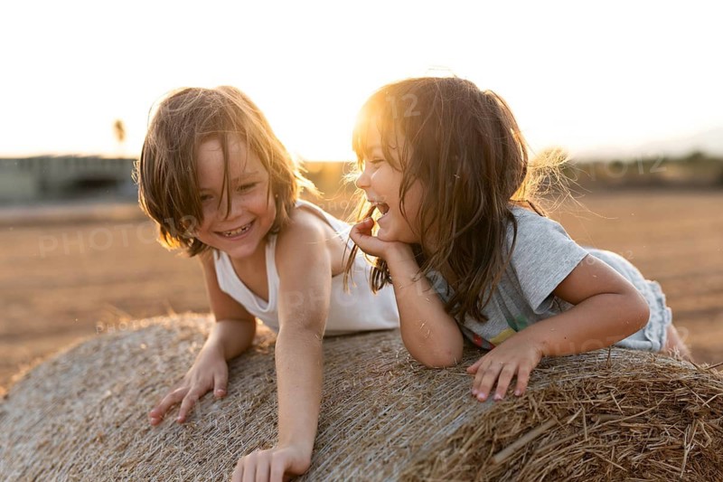 Two cheerful children are laughing and playing on a hay bale in a sunlit field, surrounded by golden, warm light. Their carefree joy captures the essence of childhood