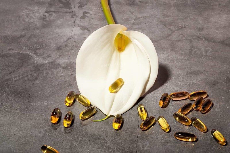 A refined composition featuring a pristine white calla lily alongside golden oil capsules on a textured gray concrete background, symbolizing beauty and wellness