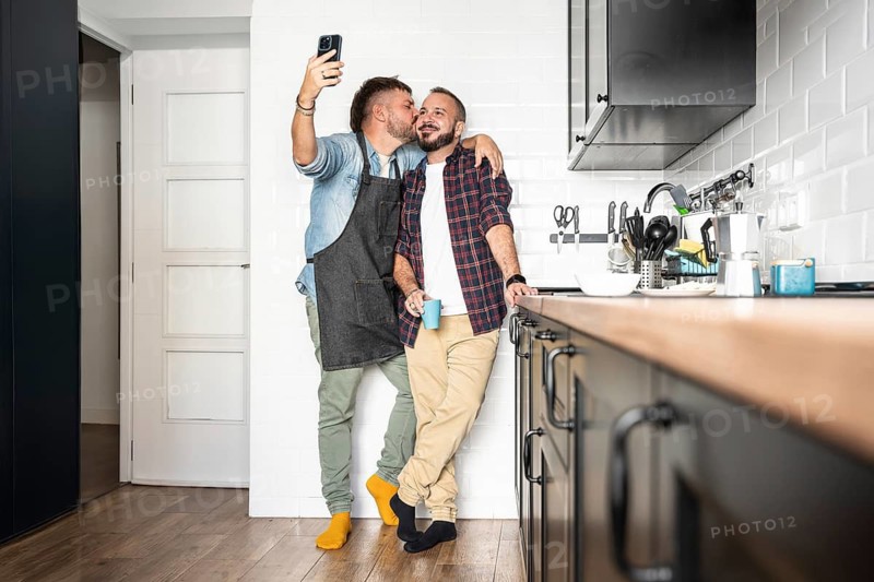A joyful gay couple poses for a selfie in their modern kitchen. One partner embraces the other, showcasing happiness and love in a cozy home setting