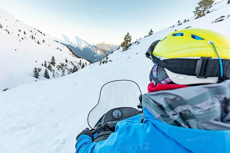A snowmobile rider in a vibrant blue jacket and yellow helmet navigates through a pristine snowy mountain landscape, surrounded by evergreen trees under a clear sky