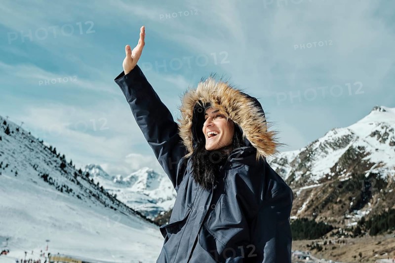 A woman joyfully waves in the snowy landscape of Astun ski resort in the Huesca Pyrenees, wearing a warm hooded coat. The majestic mountains provide a stunning backdrop