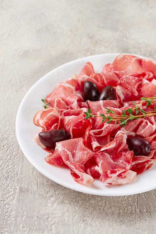 Freshly cut dried meat, served with olives and herbs, on a white plate, light background, no people