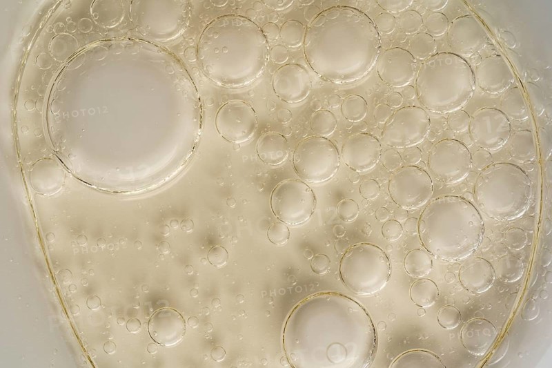 Close-up image capturing the intricate texture of oil bubbles suspended on a translucent glass surface. The macro perspective highlights the interplay of light and liquid