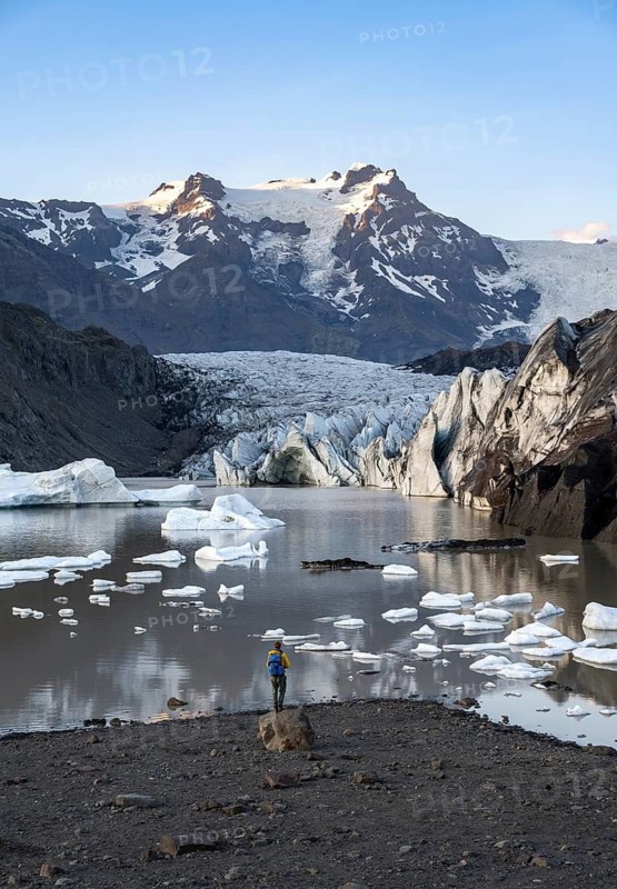 Hiker standing on a rock, reflection in the Svínafellslon glacier lagoon with ice floes, Svínasfellsjökull glacier tongue, glaciated mountain peak Hrútfjallstindar in the background, at sunset, Svínafell, Iceland